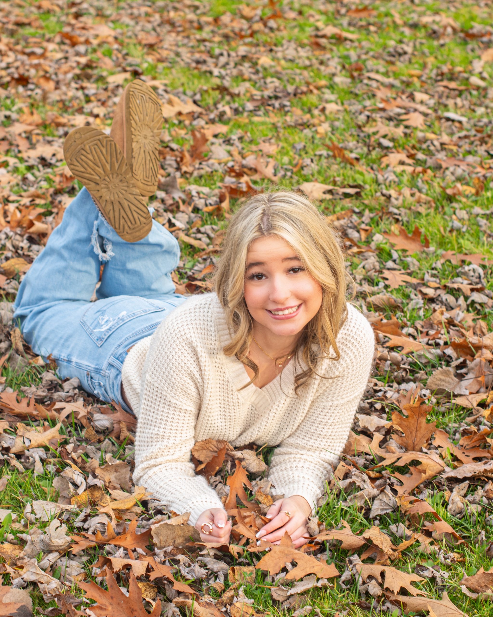 senior girl lying on autumn leaves in a forest setting, dressed in cozy sweater and jeans