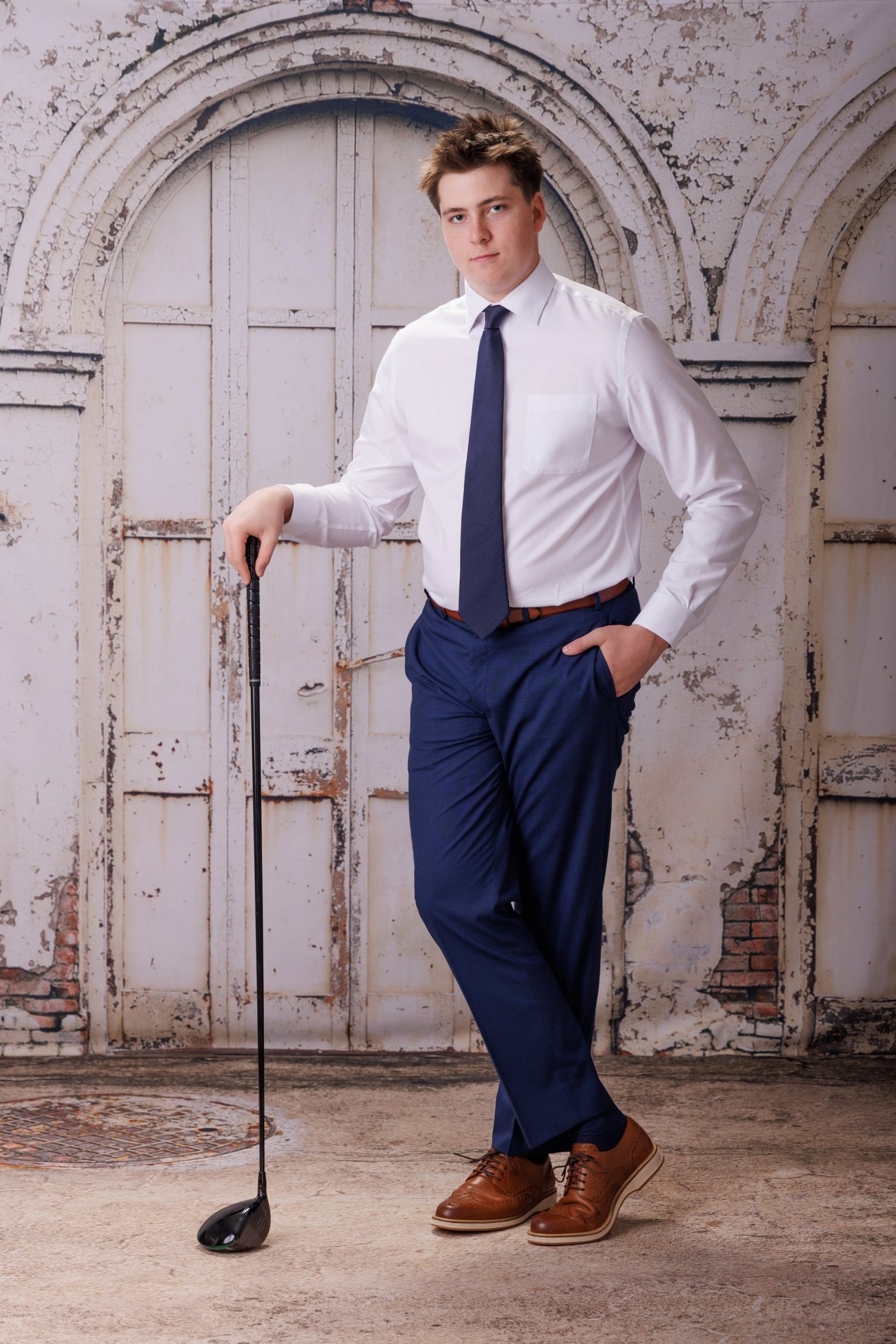 Golf enthusiast posing with club over shoulder, crisp white shirt and navy tie, vintage building facade background