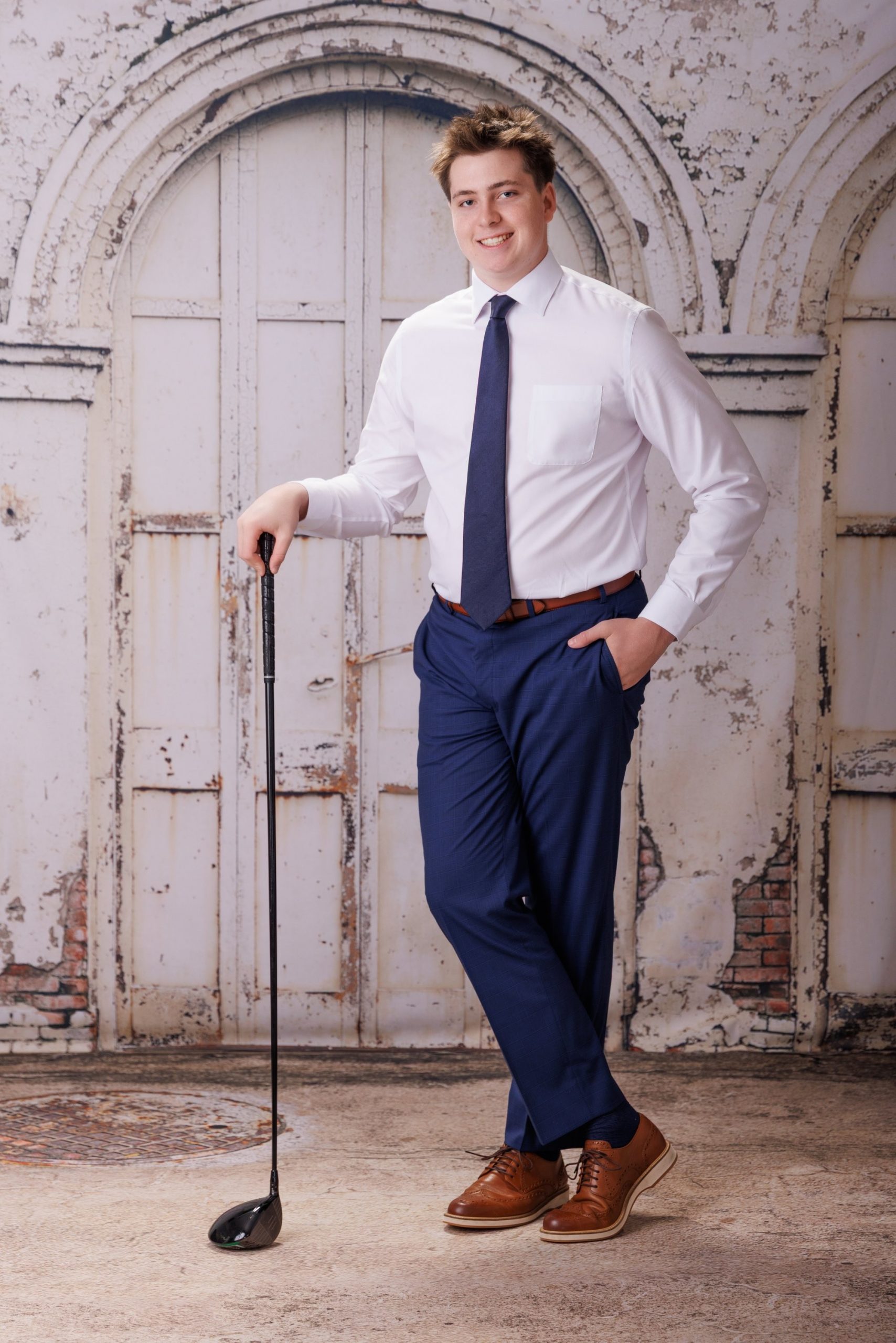 Golf enthusiast posing with club over shoulder, crisp white shirt and navy tie, vintage building facade background