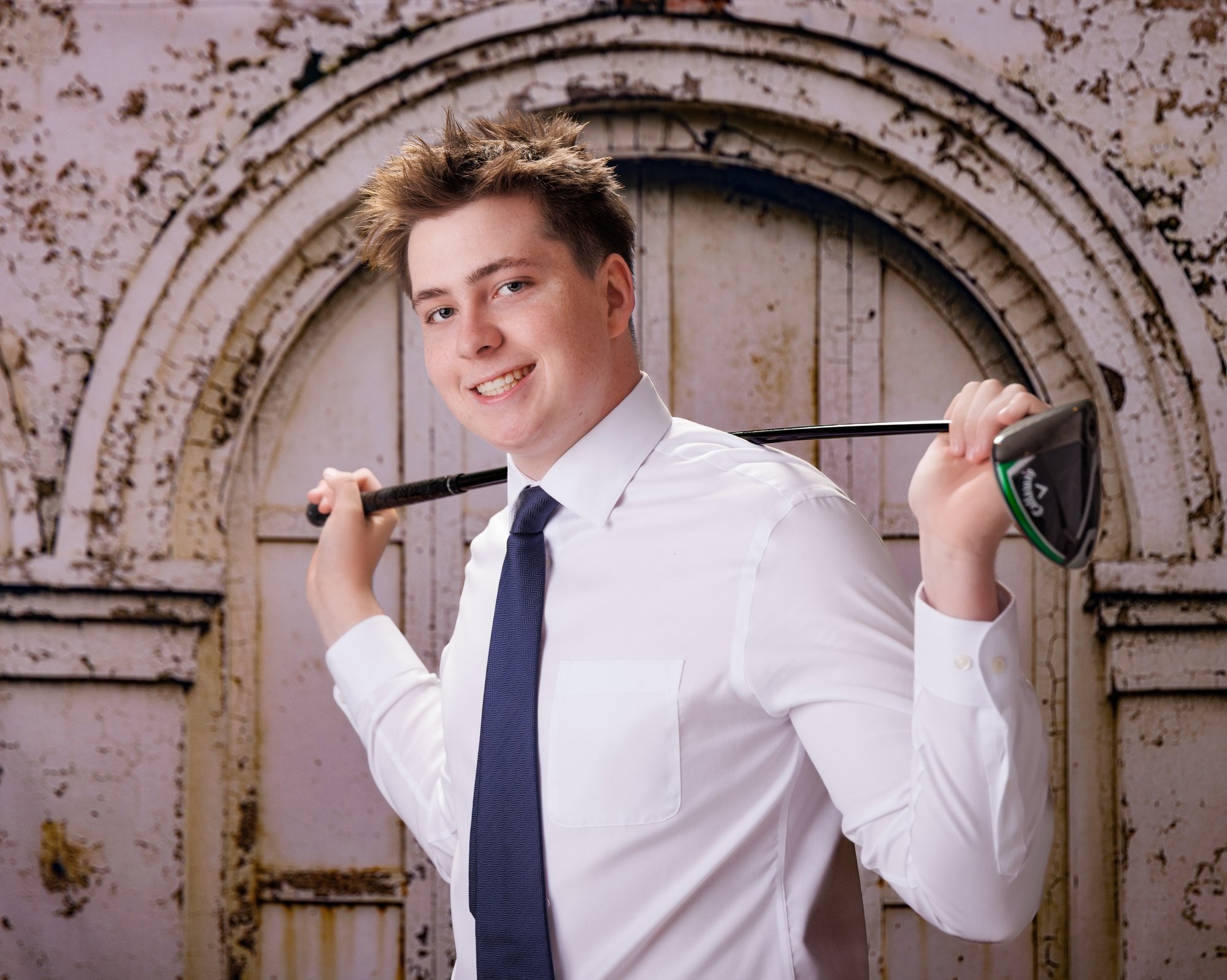 Golf enthusiast posing with club over shoulder, crisp white shirt and navy tie, vintage building facade background