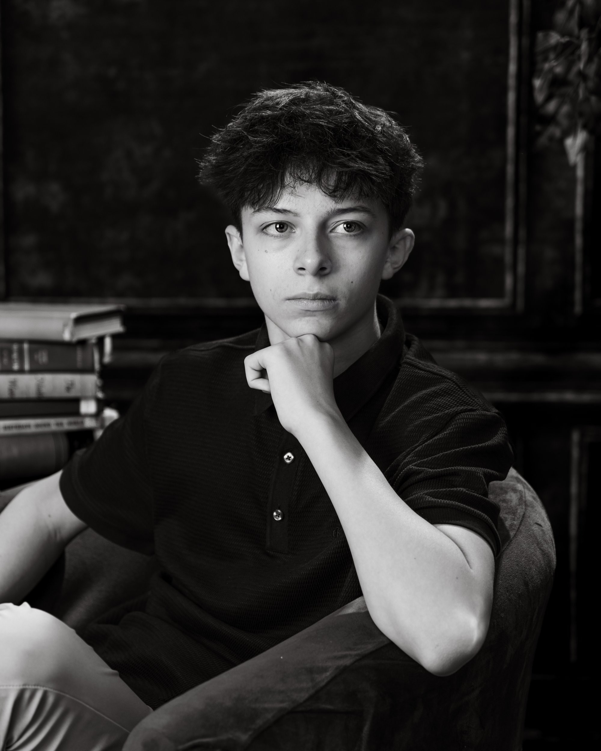 Casual senior photograph featuring a young man in a black polo shirt seated with books behind him, set in a dimly lit room