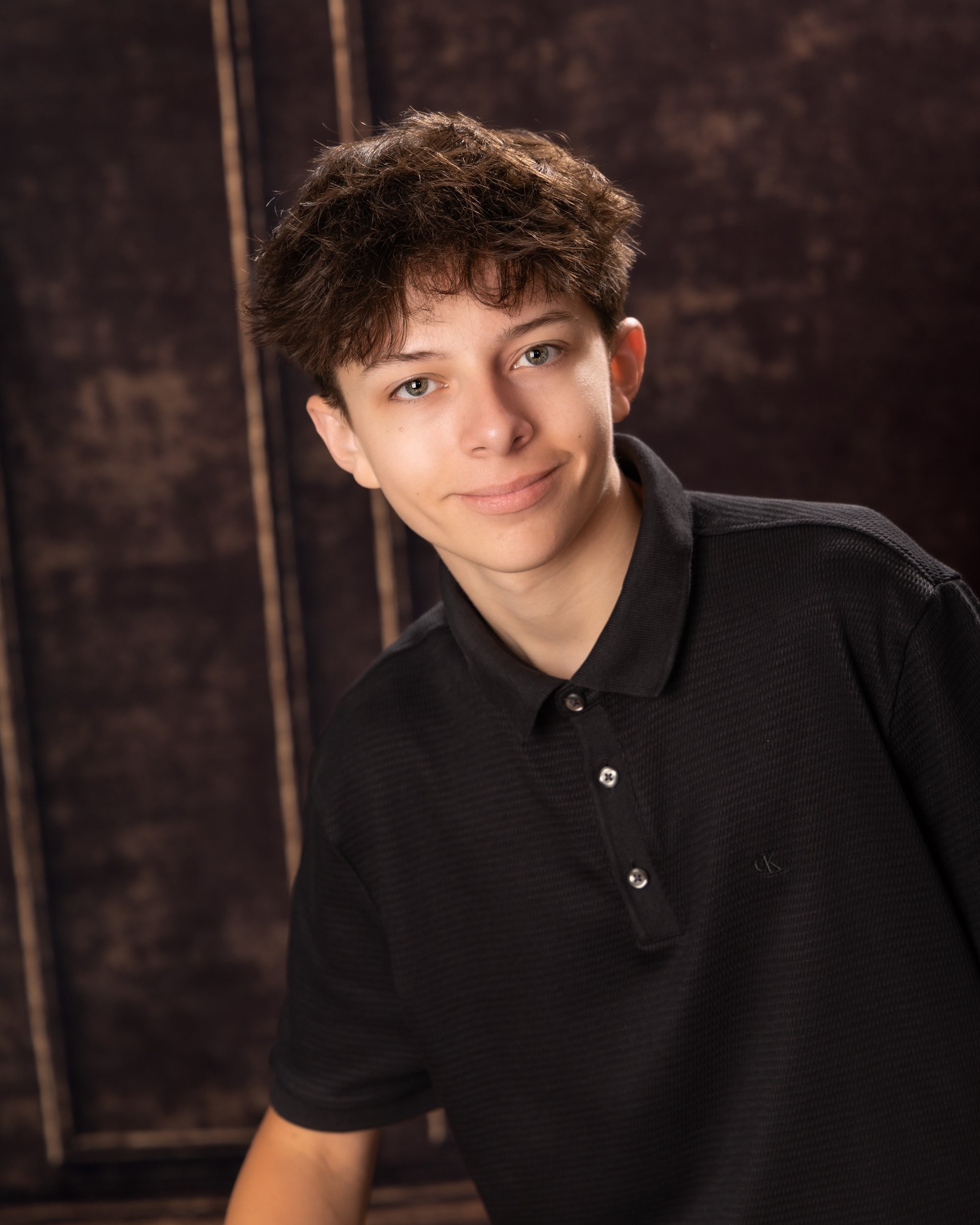 Casual senior photograph featuring a young man in a black polo shirt seated with books behind him, set in a dimly lit room
