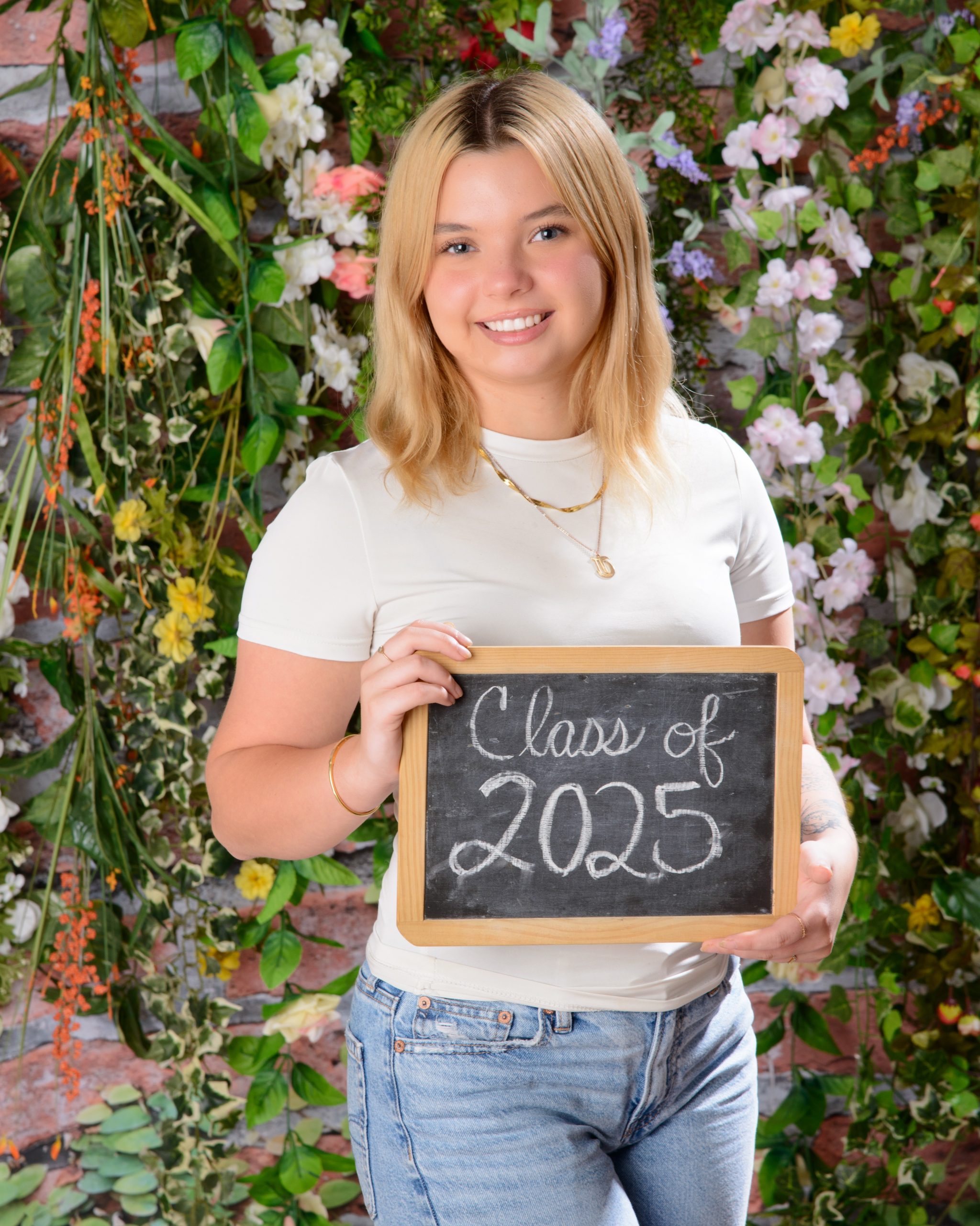 Trenton graduation photo featuring a graduate in white cap and gown holding a blue diploma cover against a lush floral background