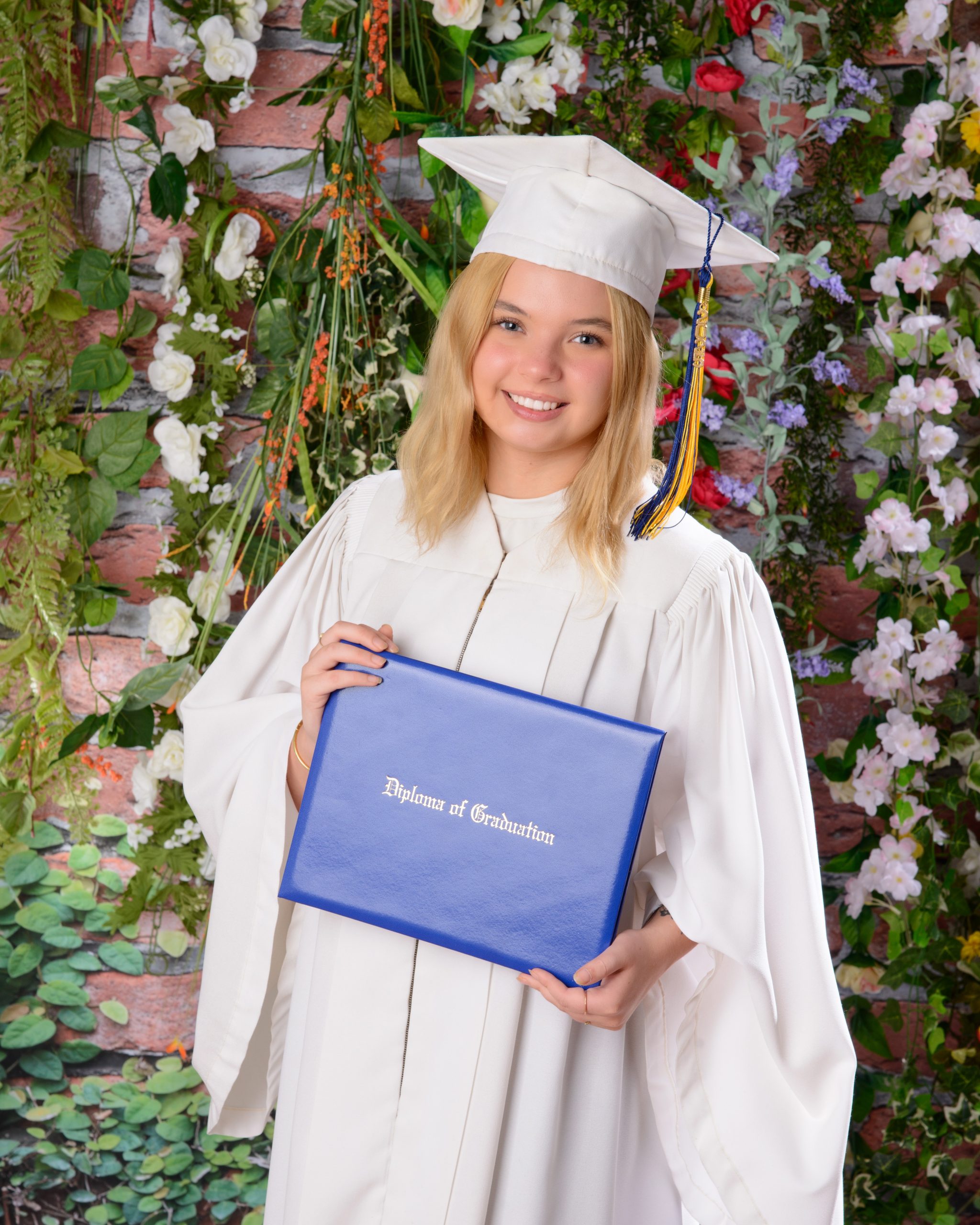 Trenton graduation photo featuring a graduate in white cap and gown holding a blue diploma cover against a lush floral background