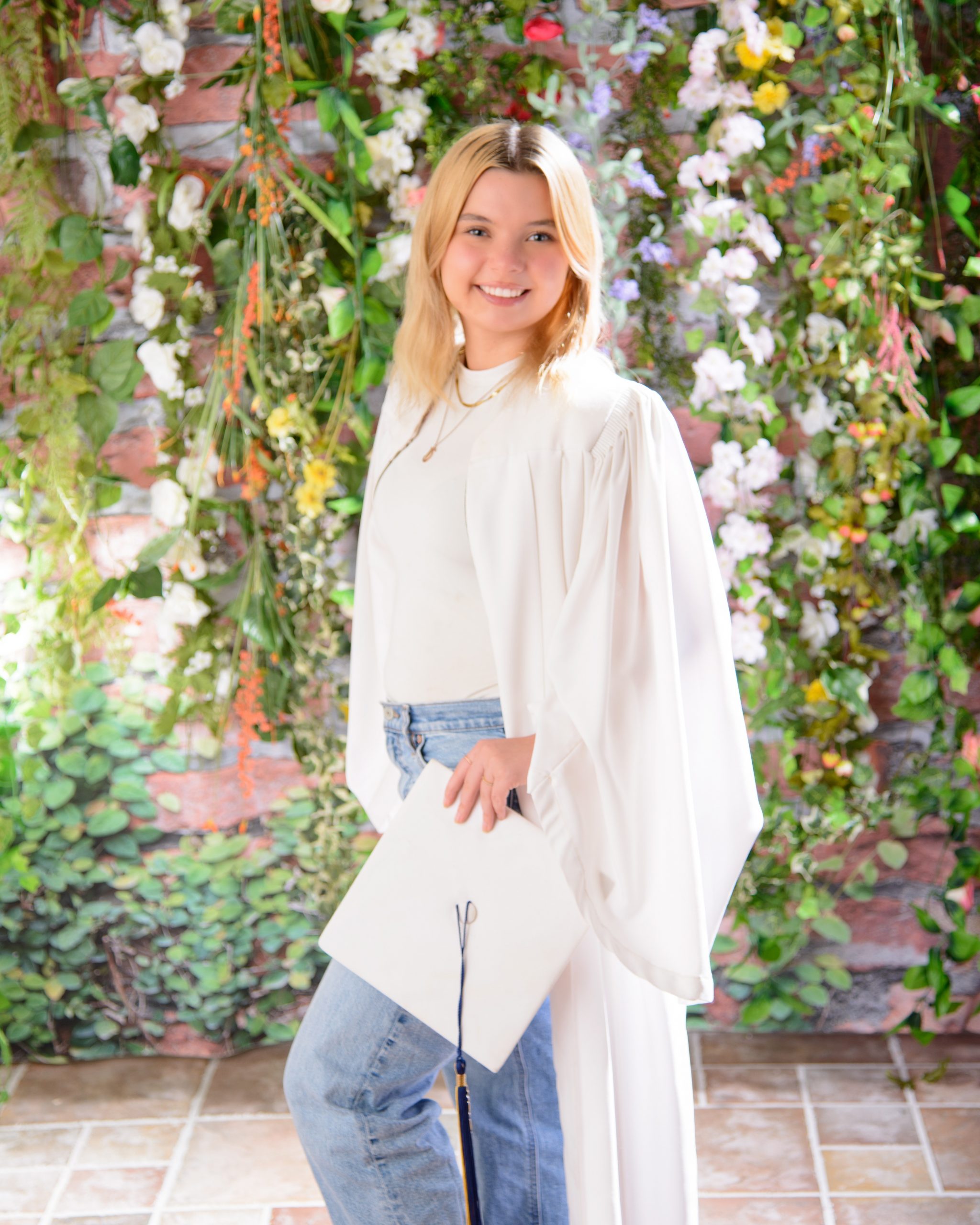 Trenton graduation photo featuring a graduate in white cap and gown holding a blue diploma cover against a lush floral background