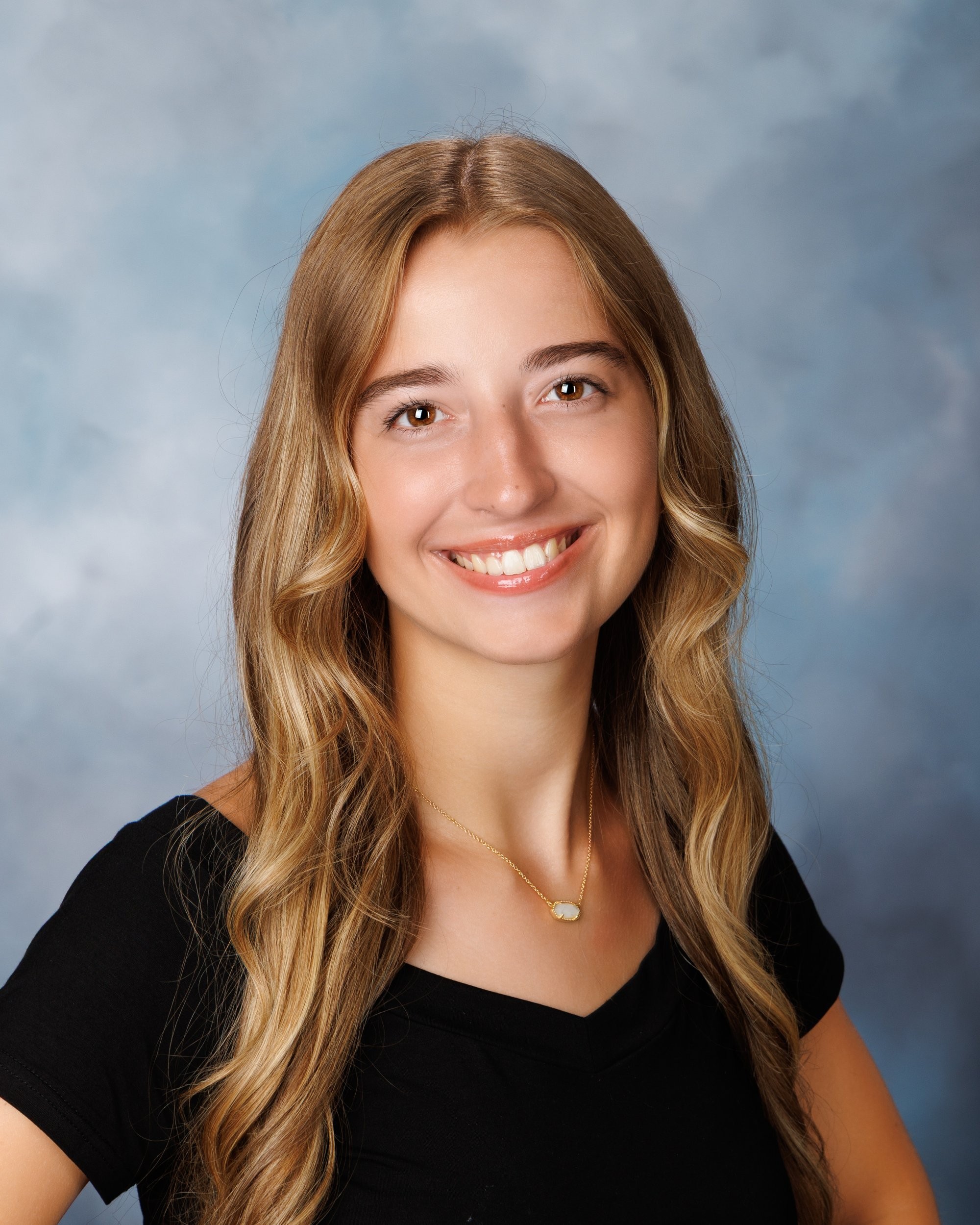Professional senior headshot of Anika with long hair, smiling against a soft blue backdrop