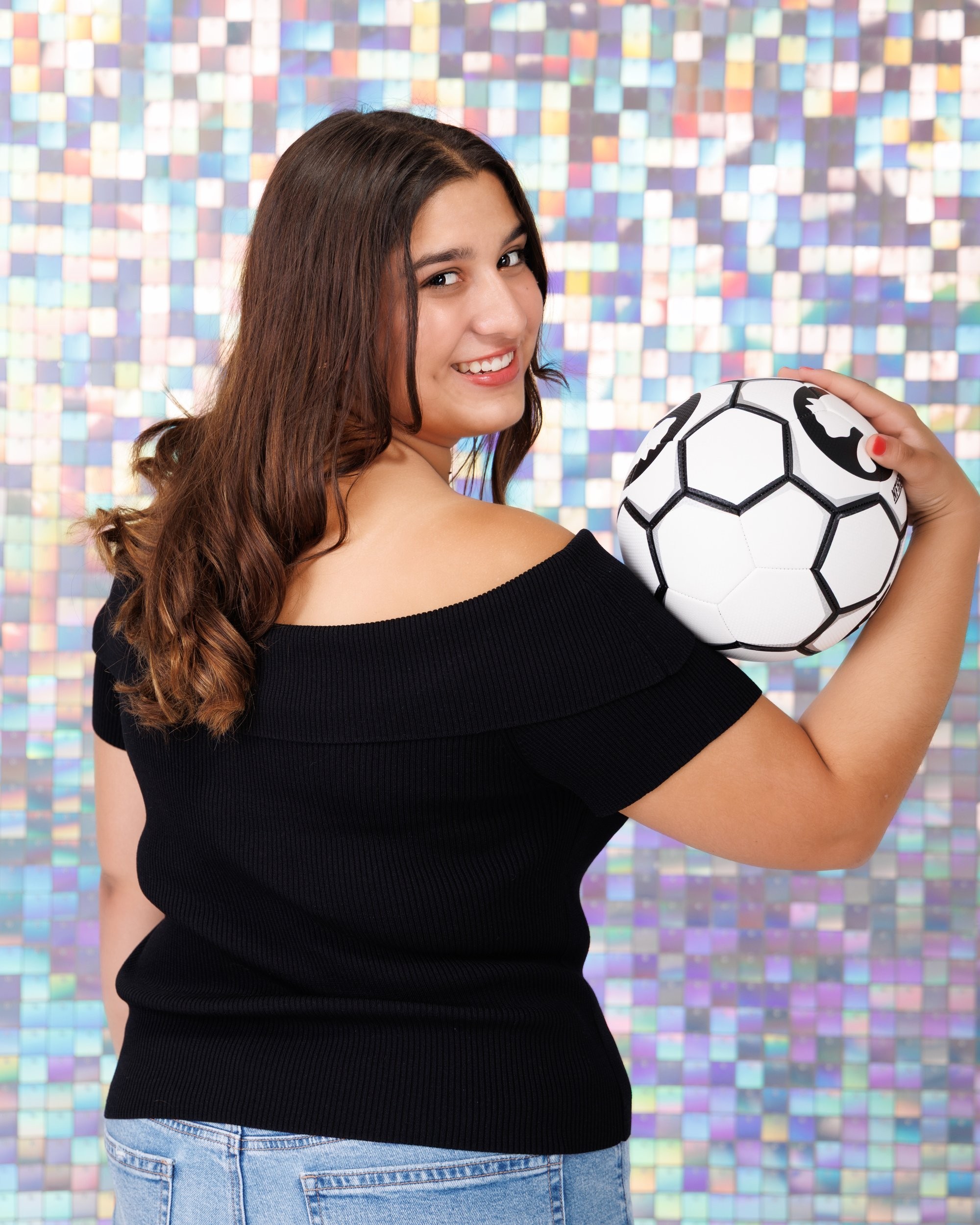 Back view of Ava holding a soccer ball in Trenton senior photo session, off-shoulder black top and mosaic wall