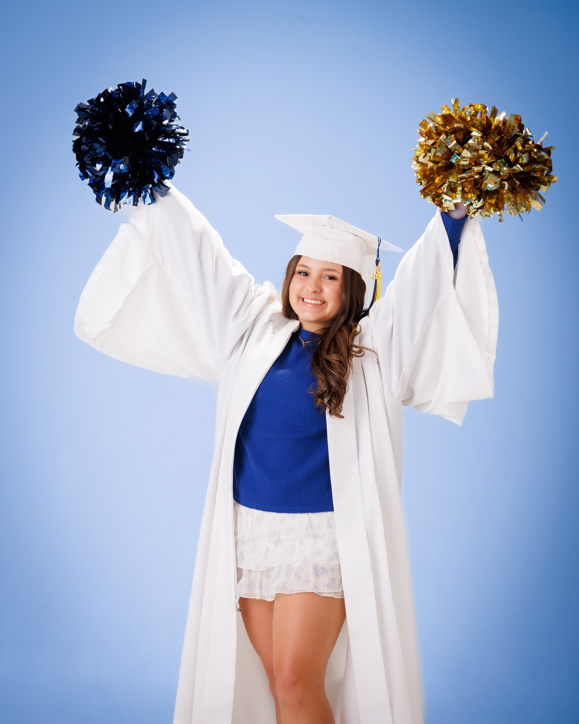 Student wearing graduation regalia with blue and gold pom-poms in a neutral backdrop for Trenton senior photography