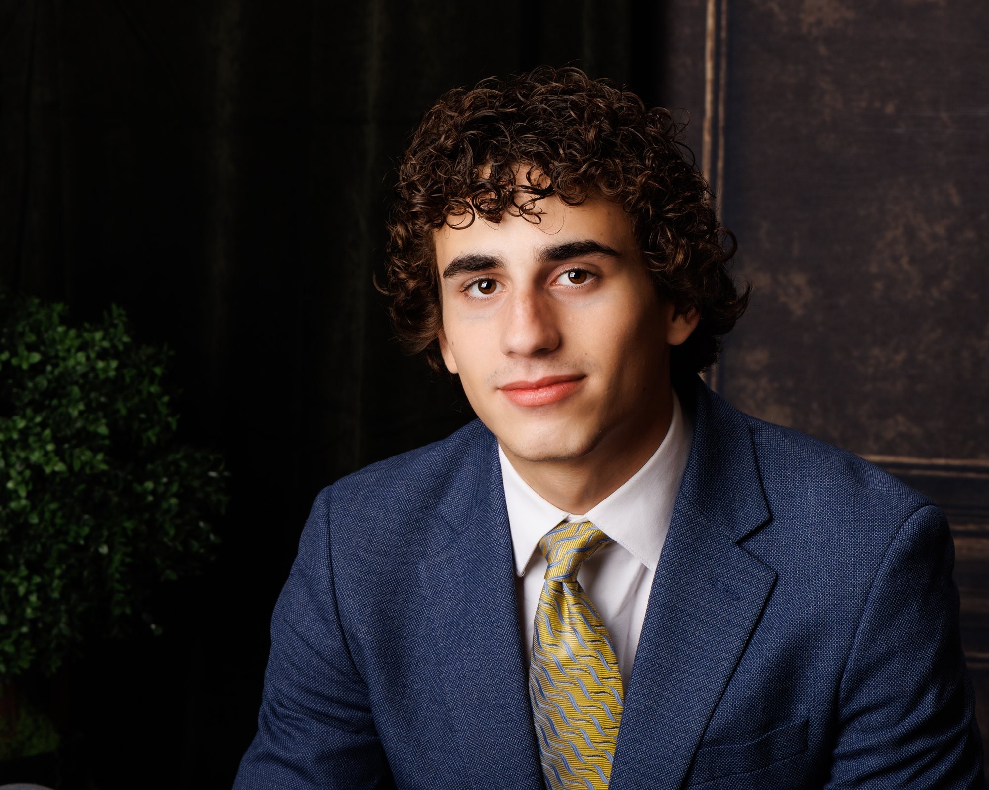 Trenton senior portrait of a young man in a blue suit, seated near greenery and a wooden wall background
