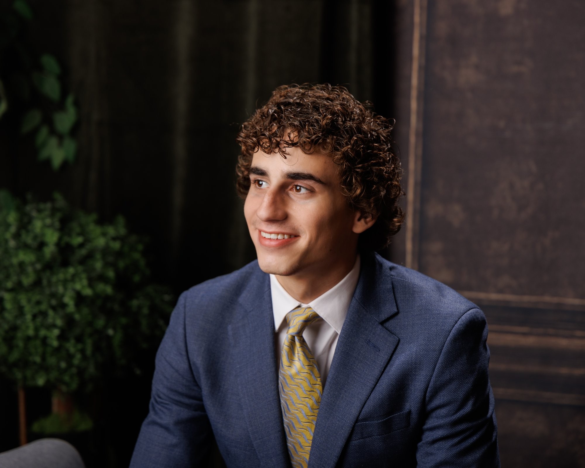 Trenton senior portrait of a young man in a blue suit, seated near greenery and a wooden wall background