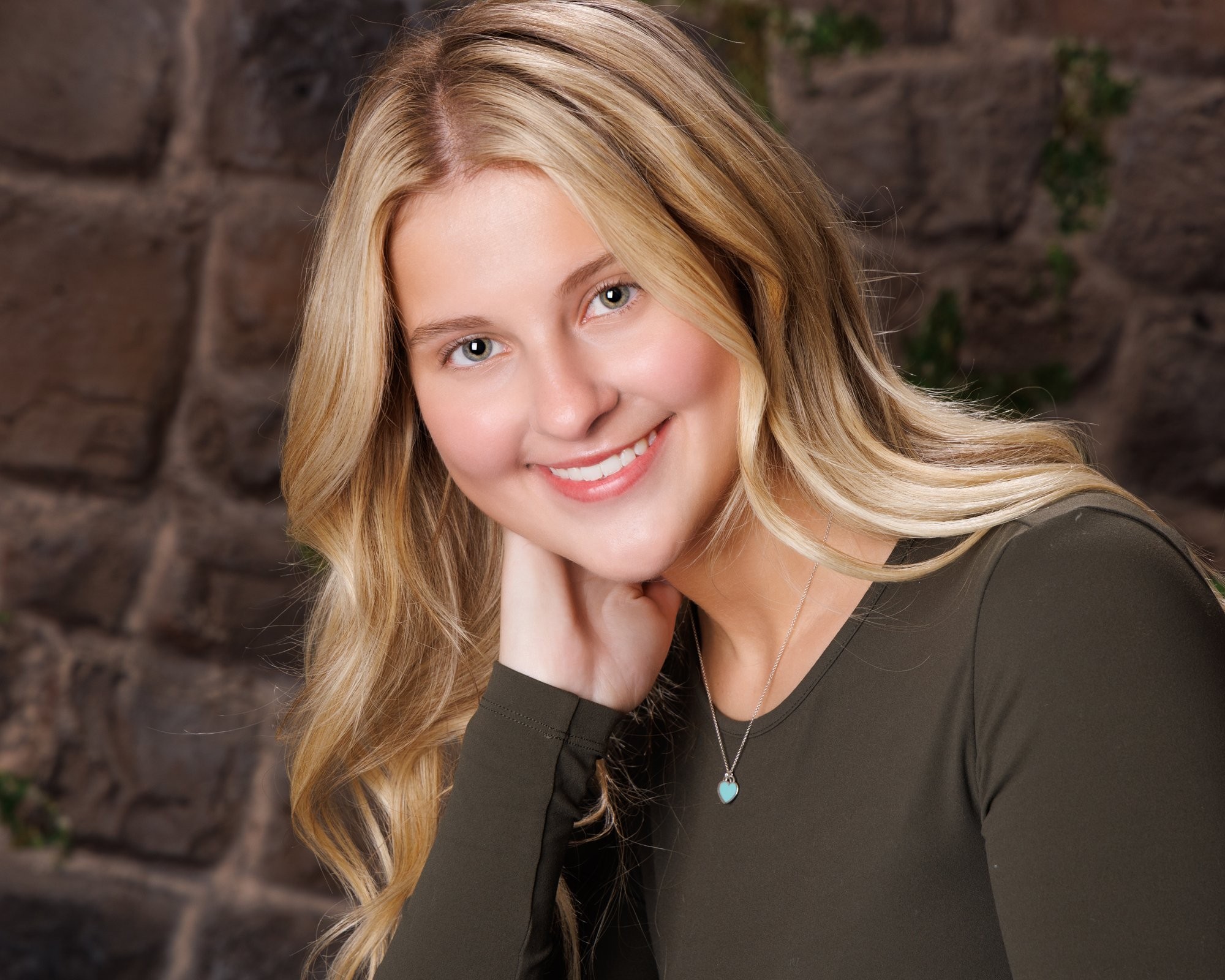 Trenton senior photography session featuring a smiling young woman with long blonde hair, wearing a green top and blue jeans, resting her hand near her neck against a stone wall background