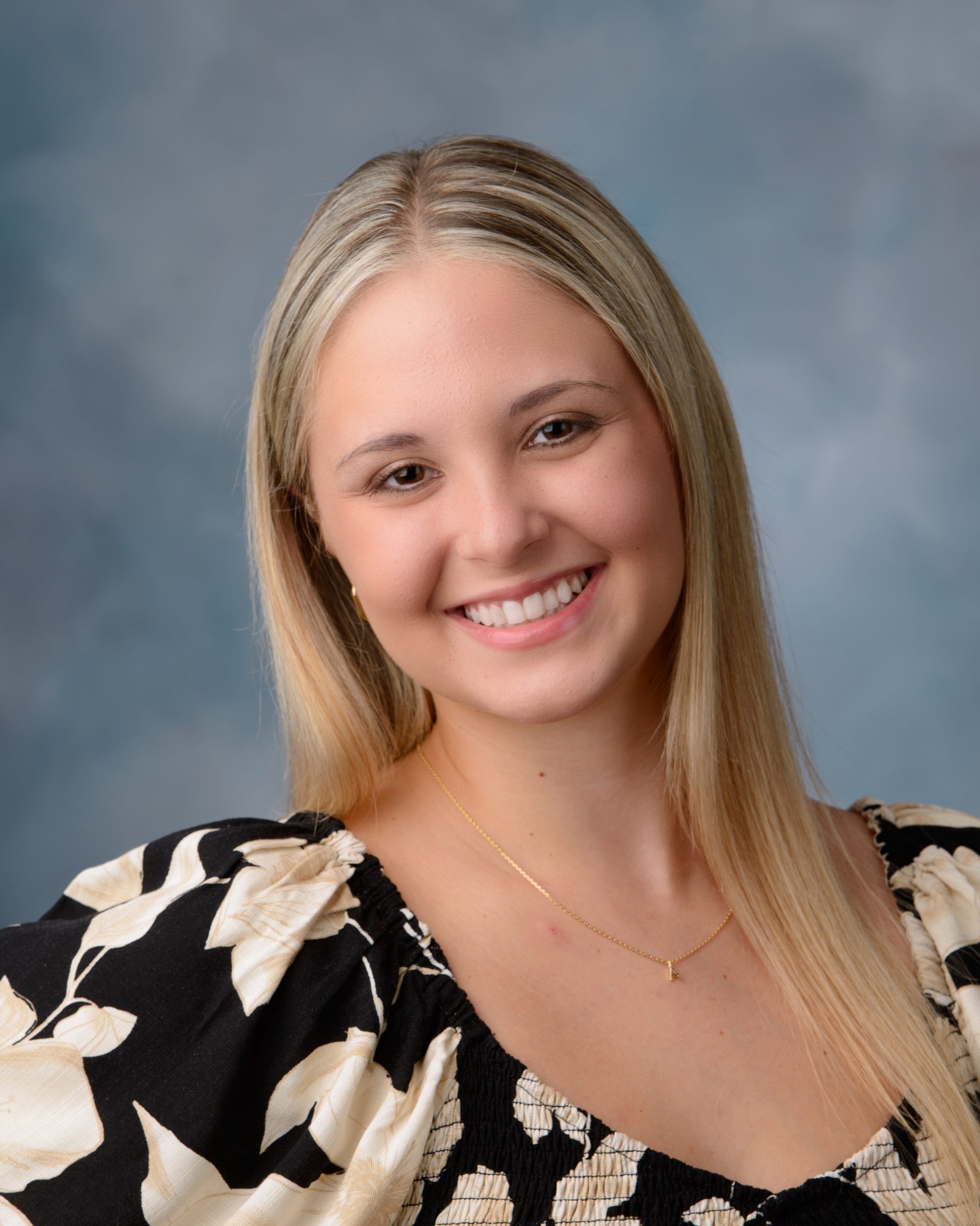Senior portrait of a woman in a black floral dress seated on a pink velvet chair, set against a pastel chevron pattern background for a vibrant Downriver photo shoot