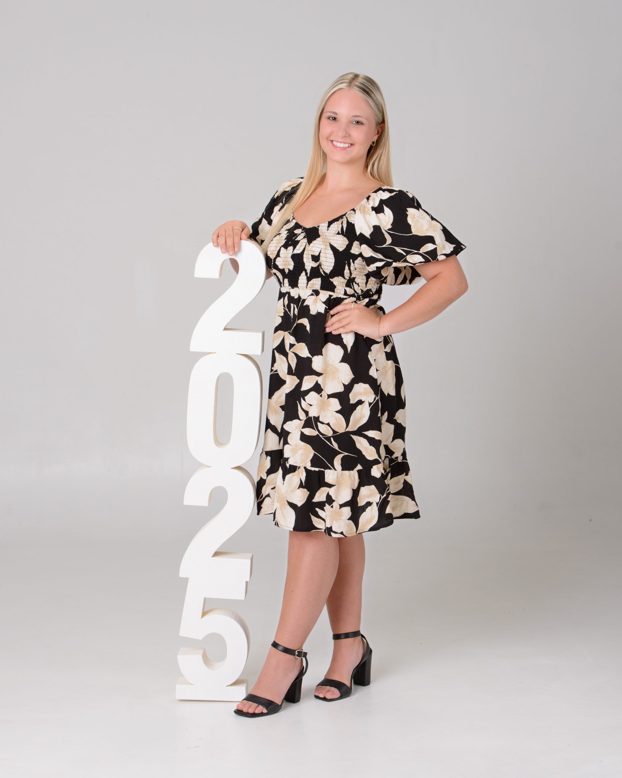 Senior portrait of a woman in a black floral dress seated on a pink velvet chair, set against a pastel chevron pattern background for a vibrant Downriver photo shoot