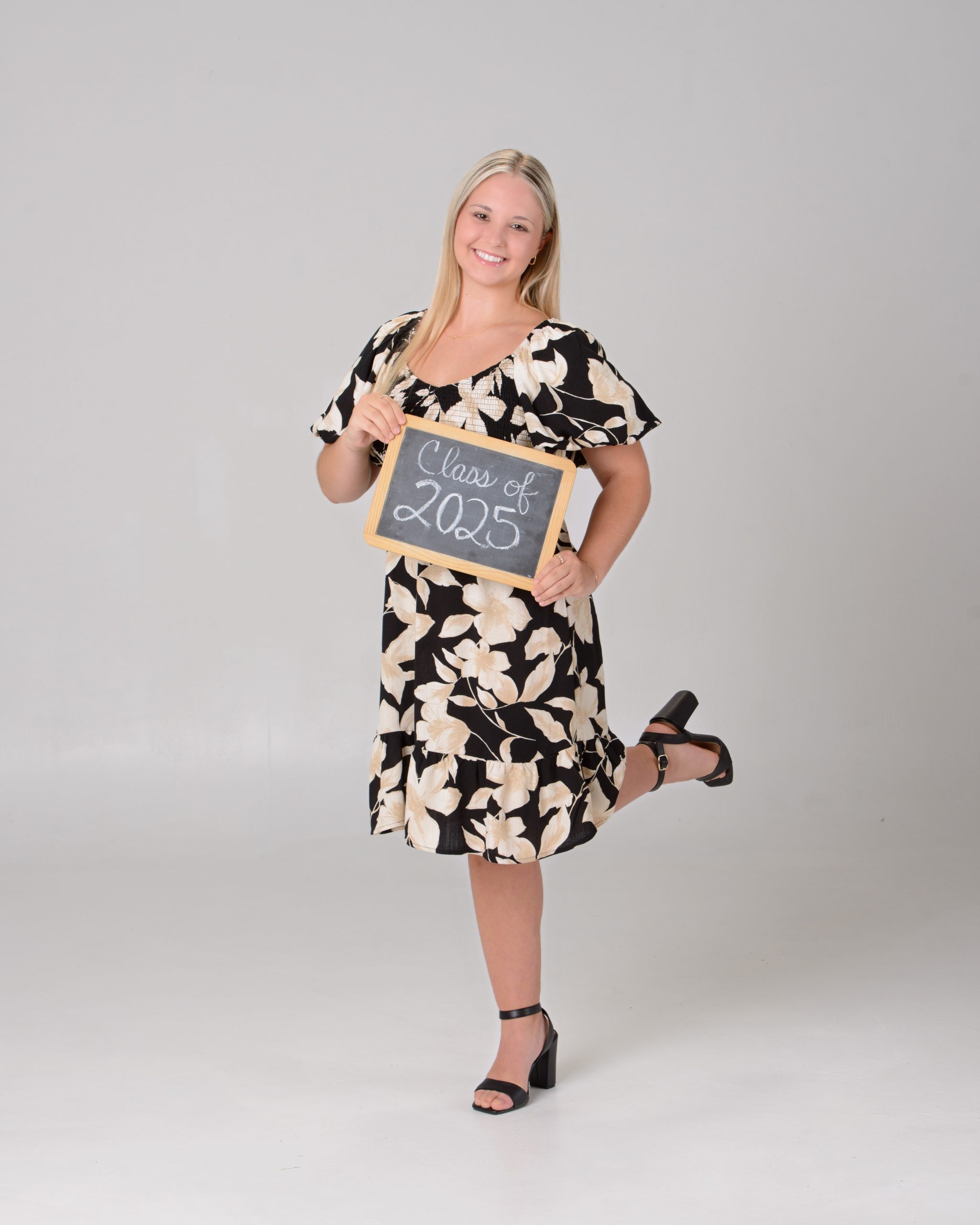 Senior portrait of a woman in a black floral dress seated on a pink velvet chair, set against a pastel chevron pattern background for a vibrant Downriver photo shoot