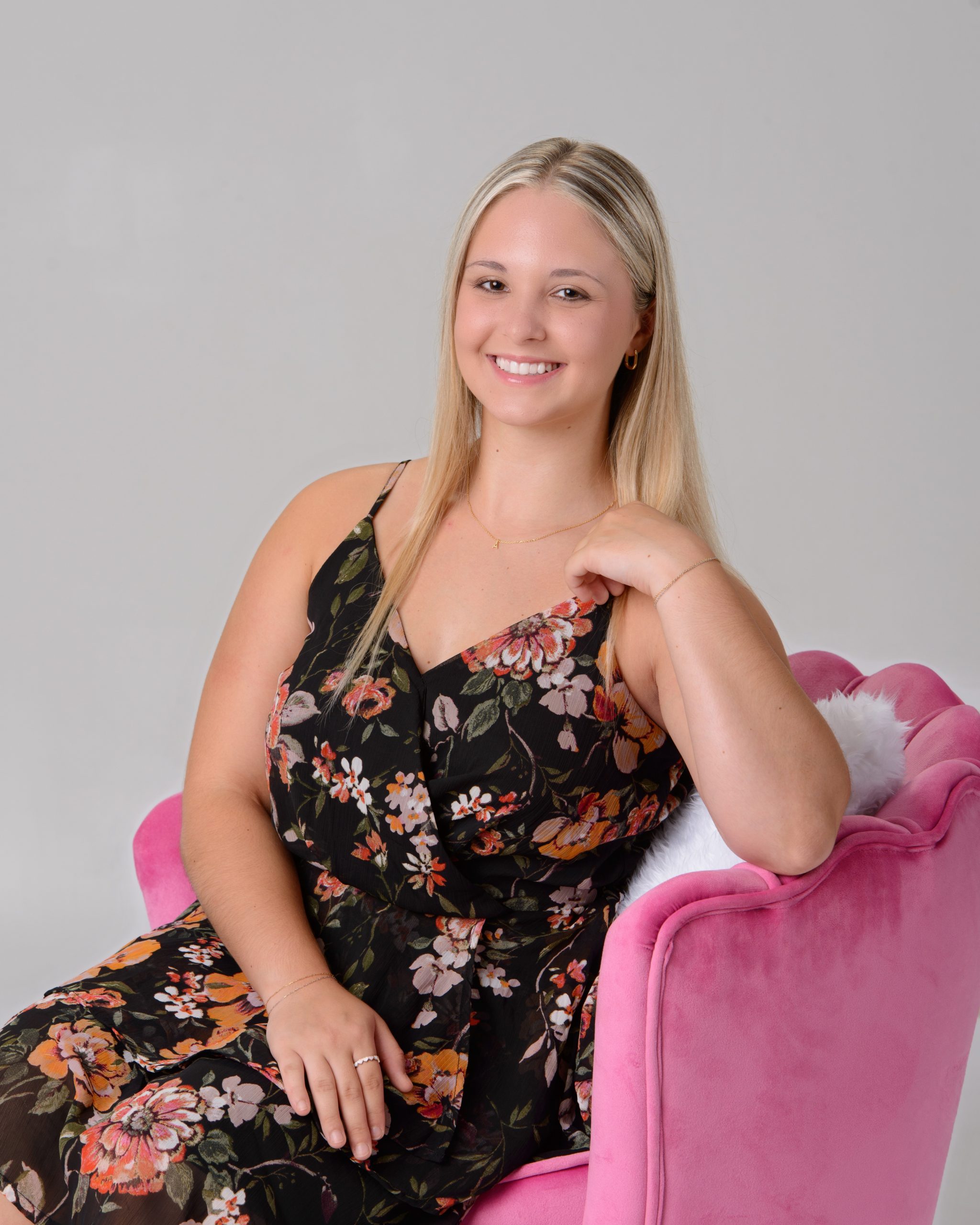 Senior portrait of a woman in a black floral dress seated on a pink velvet chair, set against a pastel chevron pattern background for a vibrant Downriver photo shoot