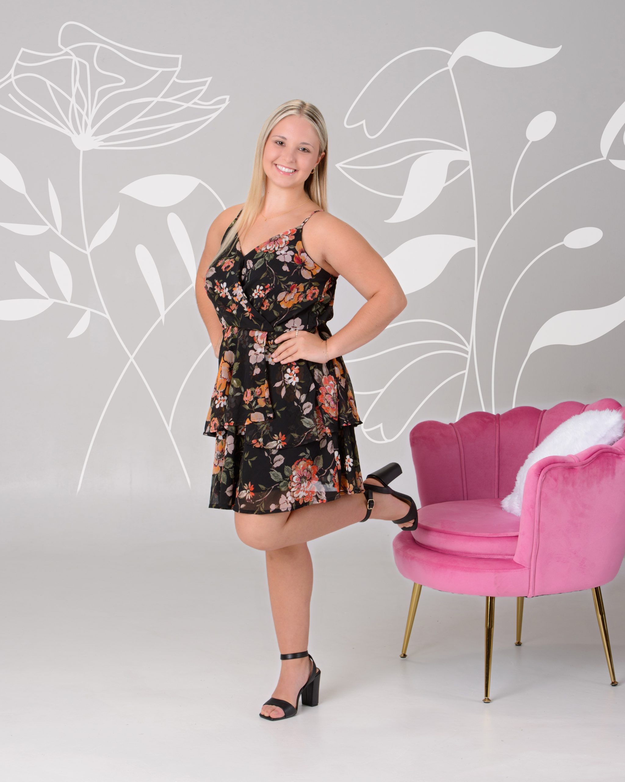 Senior portrait of a woman in a black floral dress seated on a pink velvet chair, set against a pastel chevron pattern background for a vibrant Downriver photo shoot