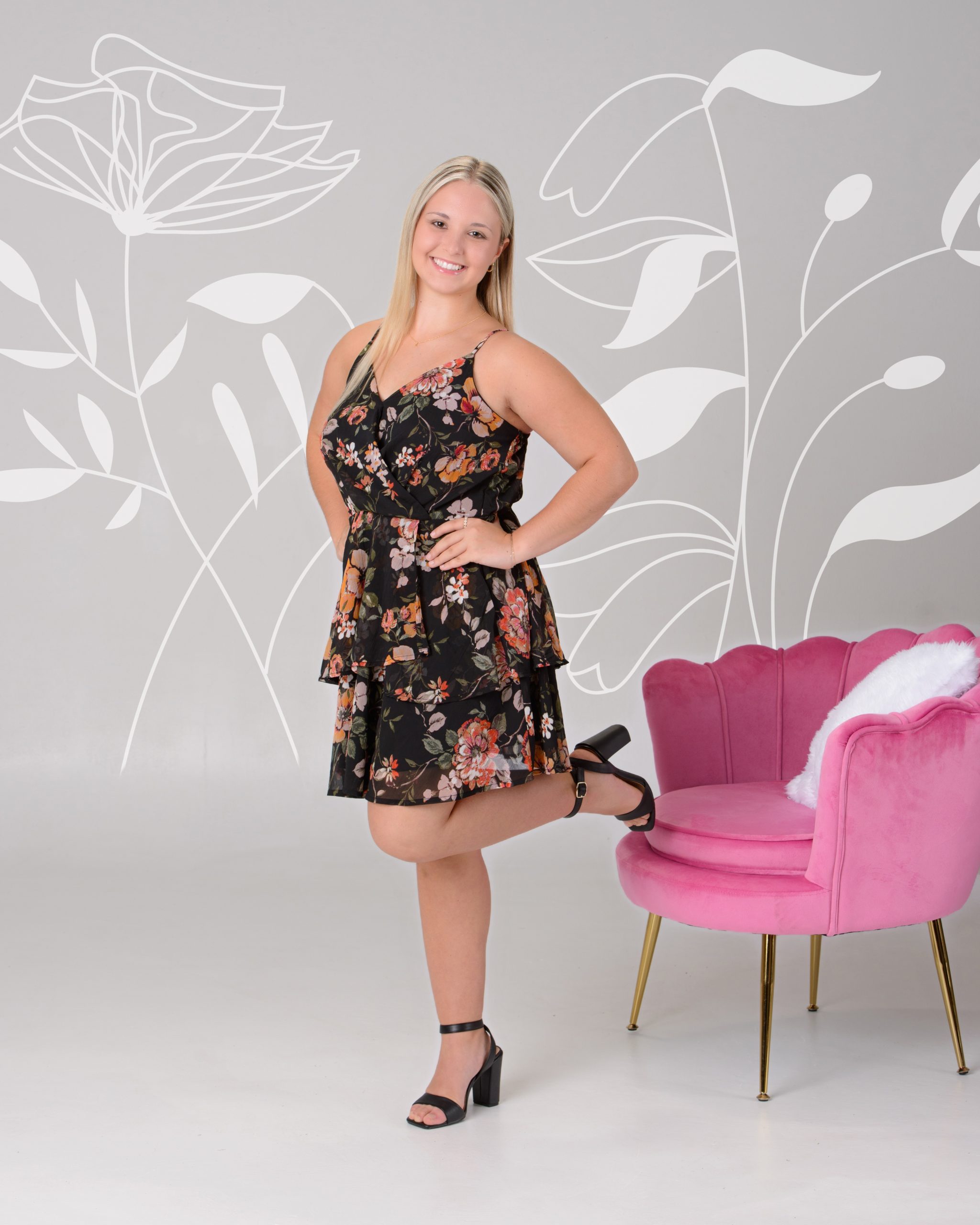 Senior portrait of a woman in a black floral dress seated on a pink velvet chair, set against a pastel chevron pattern background for a vibrant Downriver photo shoot