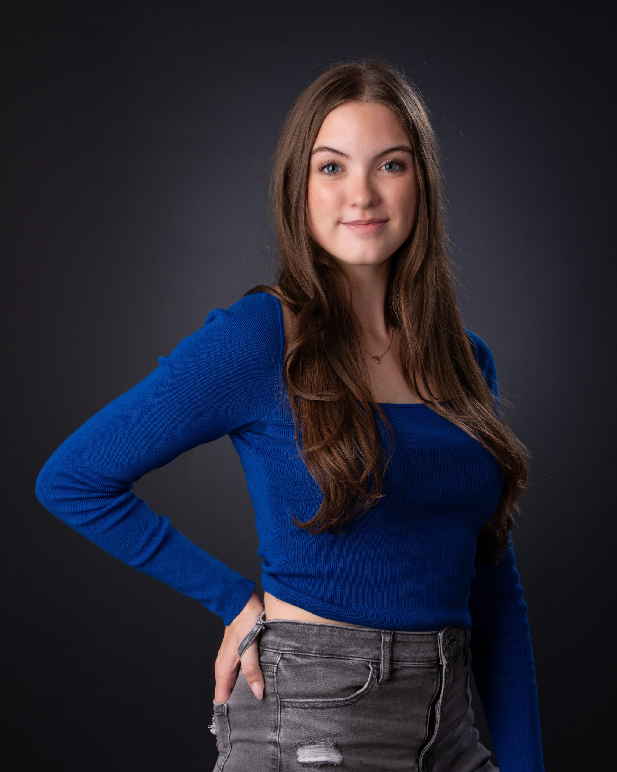 Senior girl posing with hands on hips in blue crop top and gray jeans, dark studio background