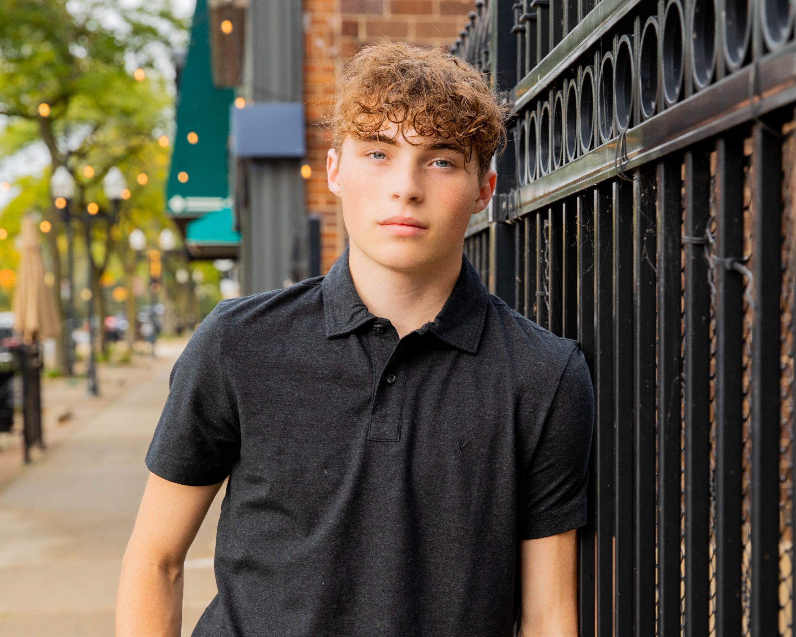 Michael in black polo shirt leaning against iron fence on urban street, Wyandotte senior photo session