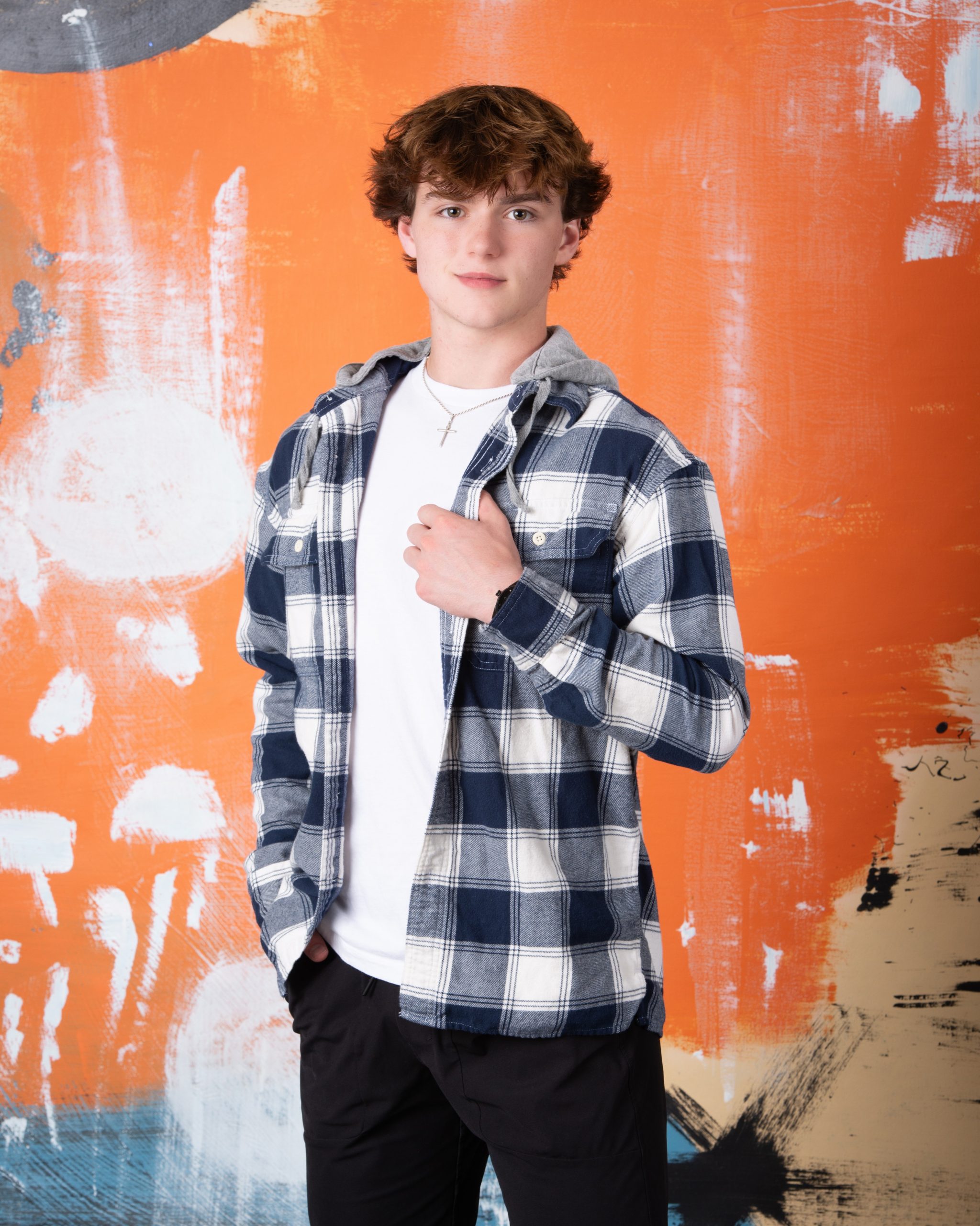 Downriver senior photography portrait of a young man seated on a wooden box in a hooded plaid jacket, white tee, black pants, and watch