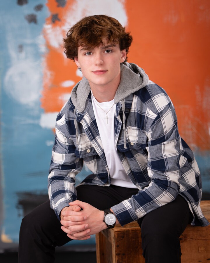 Downriver senior photography portrait of a young man seated on a wooden box in a hooded plaid jacket, white tee, black pants, and watch